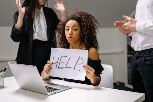 A young woman sits in front of a laptop holding up a sign asking for 'Help'
